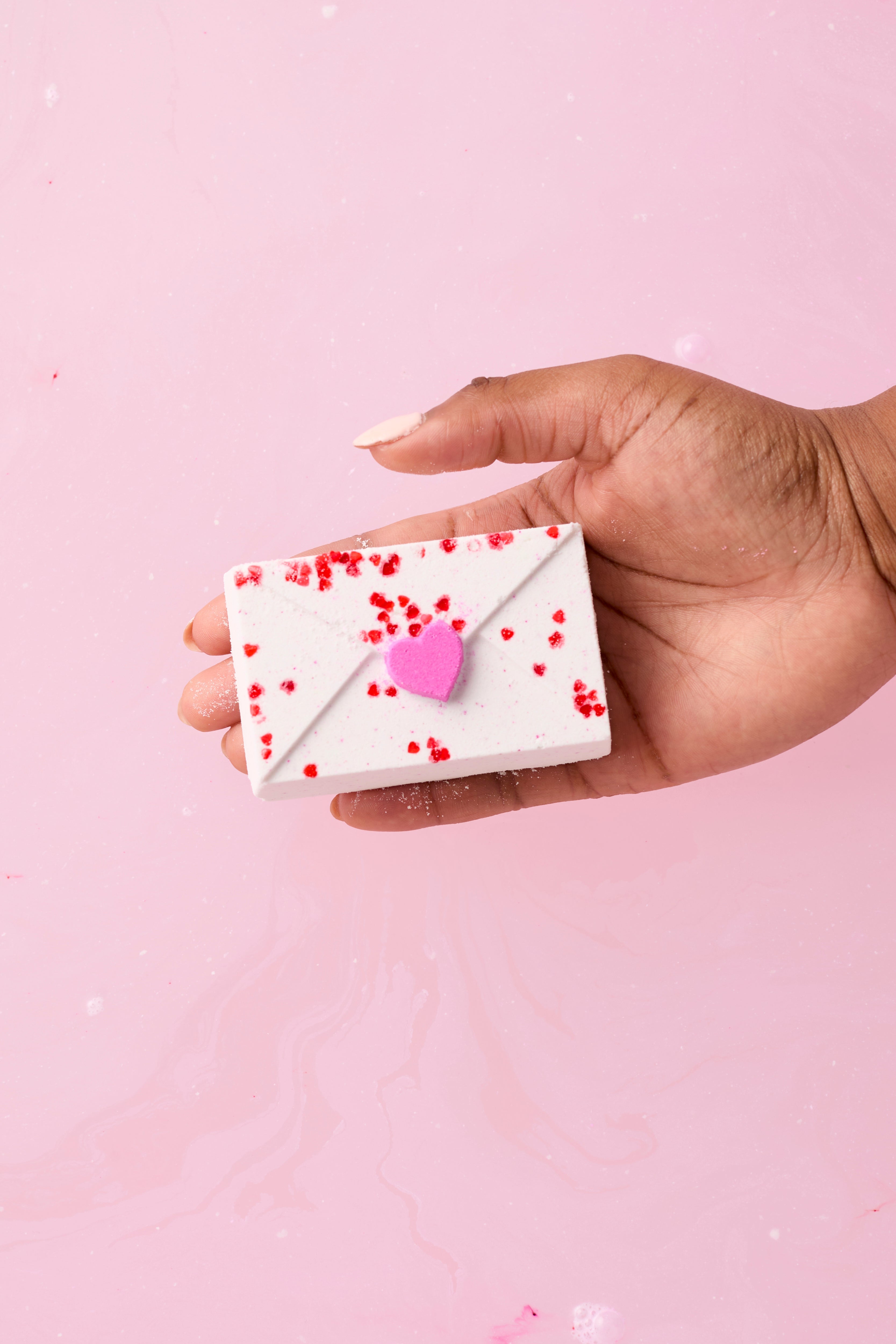Hand holding a small white bath bomb with red hearts and a pink heart on a bath