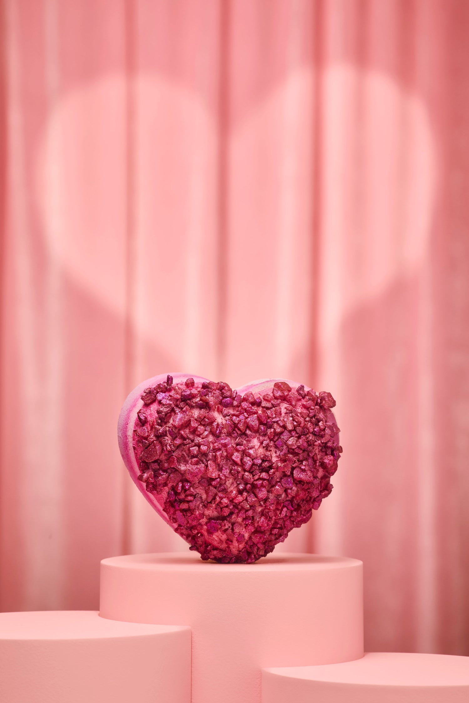 Heart-shaped pink bath bomb on a pink pedestal with a pink curtain background