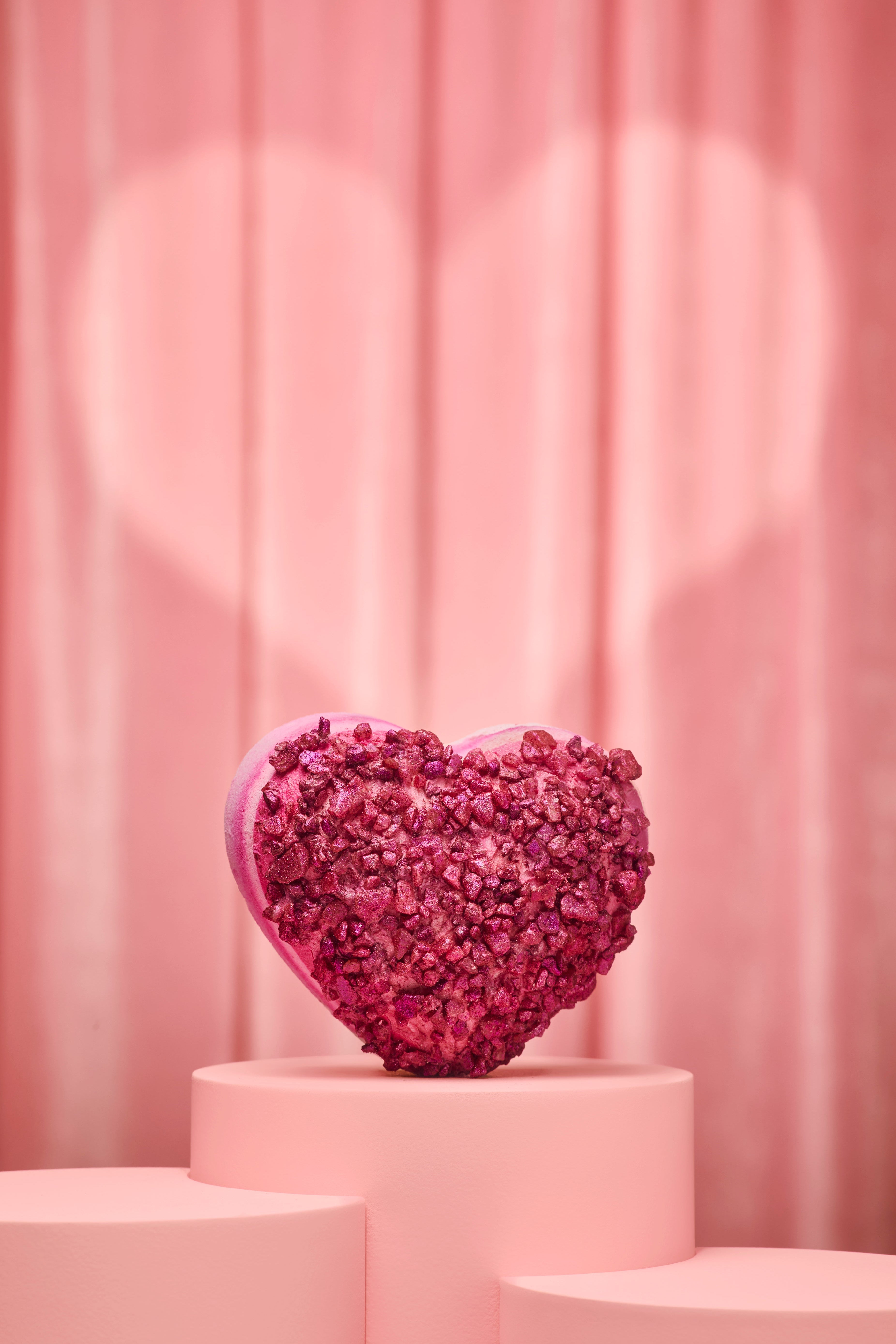 Heart-shaped pink bath bomb on a pink pedestal with a pink curtain background