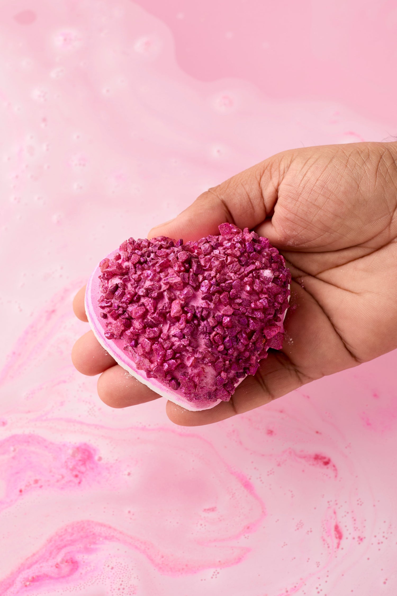 Heart-shaped pink bath bomb held in a hand with a pink bubble bath background