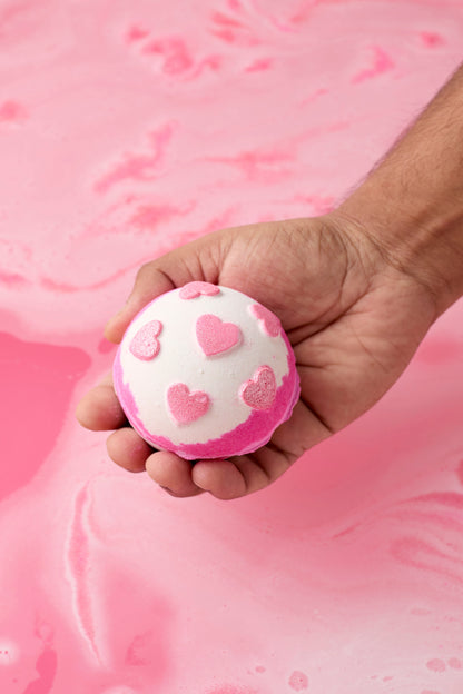 Hand holding a pink bath bomb with heart designs against a pink background